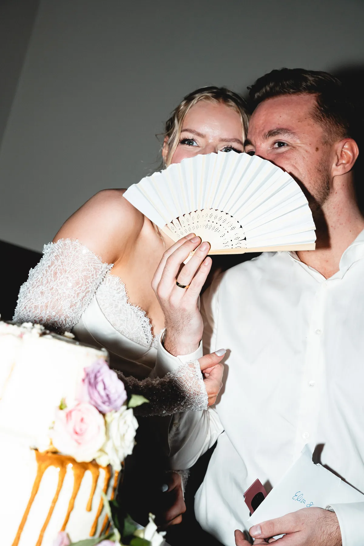 Brautpaar versteckt sich lachend hinter einem weißen Fächer, davor die Hochzeitstorte