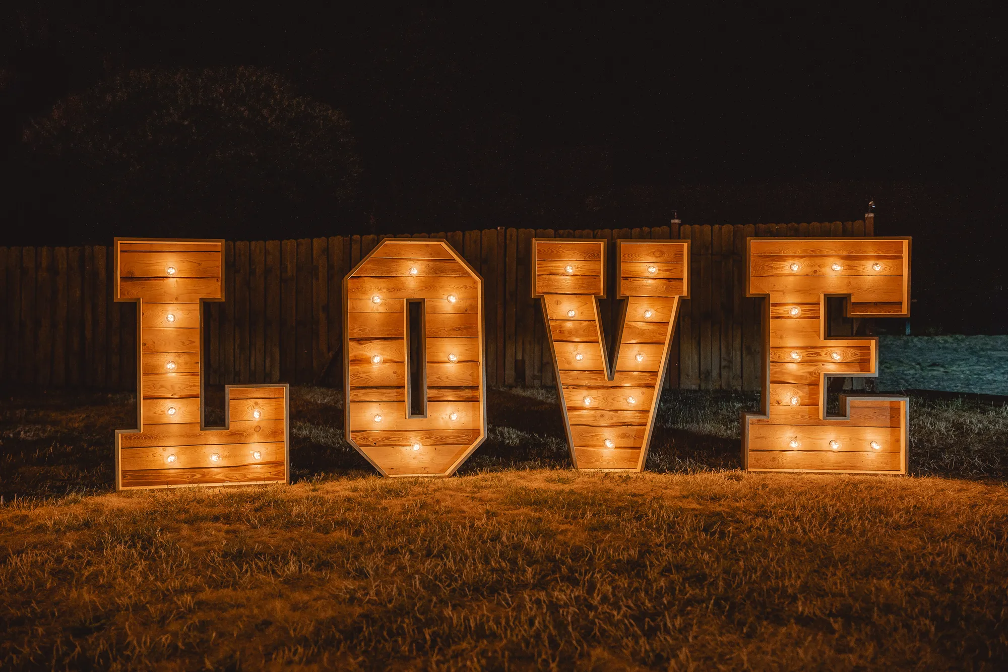 Leuchtende XXL LOVE-Buchstaben stehen nachts auf einer Wiese vor einem Holzzaun