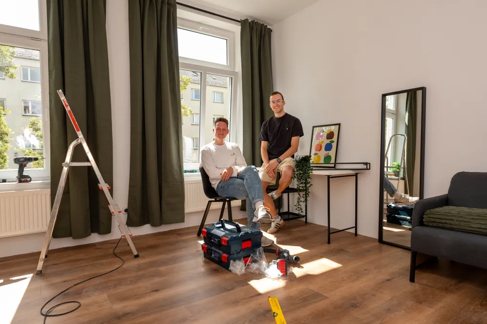 Nachher: Moritz und sein Geschäftspartner im fertig sanierten WG-Zimmer, davor Leiter und Werkzeugkoffer
